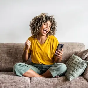 A woman sits on a couch, laughing while holding a cell phone.