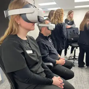 A woman wearing a virtual reality headset sits with a man in a meeting room while others stand and sit nearby.