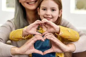 A woman and a girl making a heart shape with their hands and smiling while posing for a photograph in a room