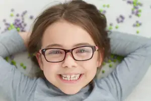 A smiling young girl with glasses lying on a white surface with colorful beads behind her