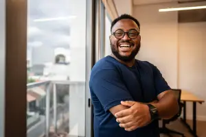 A man with a beard wearing glasses and a blue shirt is smiling and leaning against a glass window in an office room.