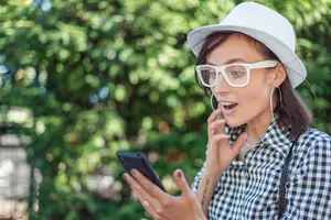 A woman wearing a white hat, glasses, and a checkered shirt is holding a cell phone in her left hand and seems to be talking to someone on the phone. She is wearing a ring on her left hand and an earring on her right ear.