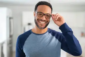 A man is smiling and posing for a picture while adjusting his glasses in a room.
