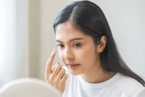 A woman with brown hair wearing a white t-shirt adjusts her contact lens in front of a mirror.