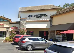 Four cars parked outside of the El Dorado Hills Optometric Center, which is a building with a stone exterior and a red umbrella in the foreground.