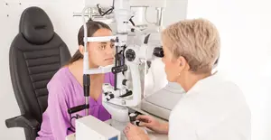 A woman is sitting on a chair and looking at a medical instrument with an older woman who is probably a doctor examining her eyes