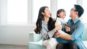 A family of three, a woman, a man, and a young girl, sitting together on a couch and laughing at something in front of them.