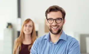 A man wearing glasses and a blue shirt smiles at the camera while a woman in a brown top stands behind him, both in an office setting.