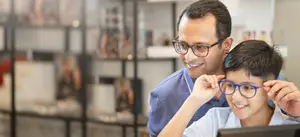 A man and a boy are standing in front of a monitor, smiling, with the boy wearing glasses and the man holding the boy's glasses.