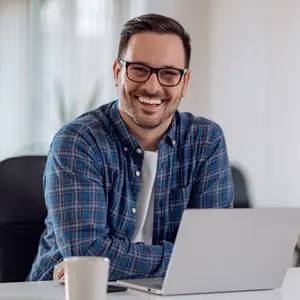 A man with glasses sitting on a chair in front of a laptop, smiling