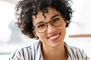 Close-up of a smiling woman with curly hair and glasses looking at the camera in a bright room