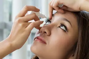 A woman applying eye drops to her eye while sitting in front of a mirror