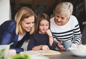A family sitting at a kitchen table, with a young girl reading a book and the adults smiling and looking at her