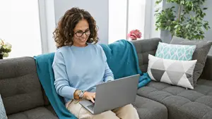 An adult woman sitting on a couch in a living room, smiling and using a laptop