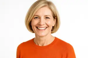 A woman with short blonde hair is wearing an orange shirt and smiling in a studio