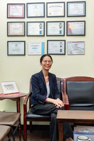 A woman wearing glasses sits in a chair in front of a wall with framed certificates in an office.