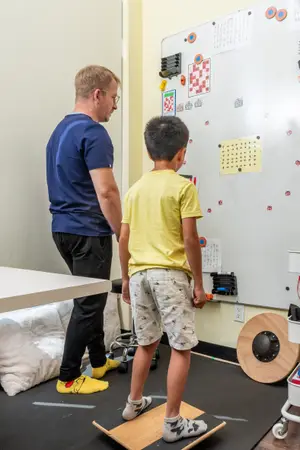 A man in glasses and yellow shoes assists a child in yellow shirt and shorts to stand on a balance board in a room with a whiteboard, cushions, and a wooden wheel on the floor.