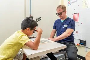 A young boy in a yellow shirt is getting an eye exam from a man in a blue shirt in an optometrist's office.