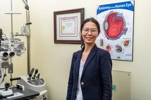 An adult woman is standing in an optometrist's office, smiling for the camera. She is wearing a navy blazer and glasses. Behind her, a framed diploma and an anatomical chart of the human eye are visible on the wall. Medical equipment, including an ophthalmoscope, is also present.