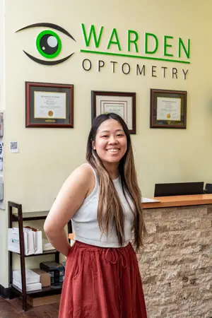 A smiling woman standing in front of a reception desk with an optometrist logo on the wall behind her