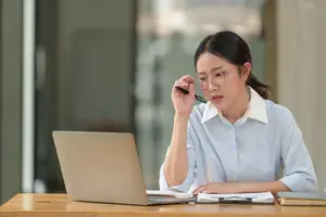 A woman wearing glasses is sitting on a chair in front of a desk, working on a laptop, and writing something in a notebook