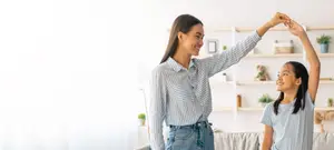 A woman and a girl are playing in a living room, they are holding hands and smiling. The woman is wearing a striped shirt and the girl is wearing a gray shirt. There is a white shelf with a basket, a potted plant, and a stuffed animal behind them. There is a white curtain on the left side and a couch on the right side.