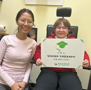 A woman wearing glasses and a young boy in a wheelchair both smiling and holding a sign that says I'm a Vision Therapy Graduate!