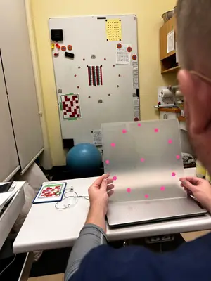 A person in a blue shirt is using a laptop with a clear plastic sheet with pink dots on it on a desk in a room with yellow walls and cabinets.