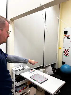 A man wearing glasses and a blue shirt is pointing at a ping pong ball attached to a string hanging from the ceiling.