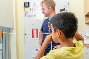 A boy with glasses looks at a wall while a man in a blue shirt watches. He holds a stick with colored beads on it.