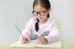 A young girl is sitting on a chair while wearing eyeglasses and writing on a notebook with a pencil