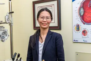 A smiling woman wearing glasses stands in front of an eye examination setup in a clinic