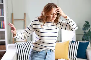 A woman is standing in a room and holding her head with her left hand while closing her eyes, probably feeling unwell.