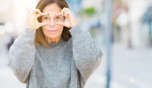 A woman in a gray sweater is making a heart shape with her hands on her forehead, looking in front and posing for a photo in a street.