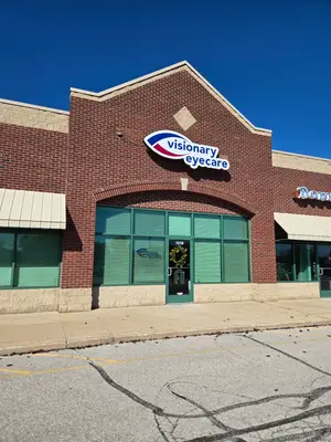 A brick building with a glass door and windows, a sign above the door reads 'Visionary Eyecare', and a wreath is placed in front of the door.