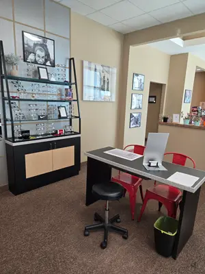 A well-lit room with a desk, red chairs, a trash bin, a shelf with glasses and a vase, and picture frames on the wall.