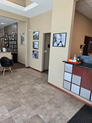 A photo of an interior space featuring a tiled floor, a wooden desk with a pumpkin, a chair, and a wall adorned with picture frames, including one with a woman's photo, is shown. The room has a ceiling with a white light, a shelf with various items, and a door at the end of the hallway. On the left side, there is a shelf with items, and on the right side, there is a wall with picture frames. The image has a watermark on the bottom right corner.