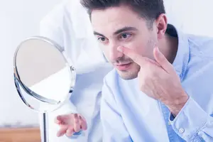 A man is examining his skin in a mirror while a medical professional stands behind him.