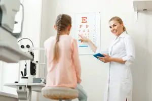 A doctor in a white lab coat points to an eye chart while a young girl looks on in an eye clinic.