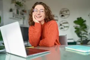 A woman in a red sweater sits at a desk with a laptop and a notebook, smiling at the camera.