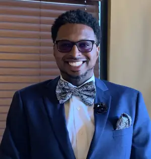 Man in a blue suit with a bow tie and glasses smiling at the camera