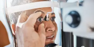 A woman is undergoing an eye examination, using an ophthalmoscope, while seated in front of a white wall with an open door, her face illuminated by a light.