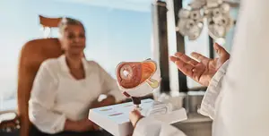 A person in a white lab coat is demonstrating an eye model to a seated woman in a hospital room.