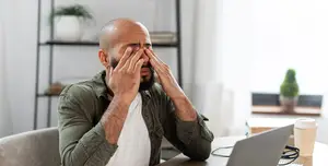A man sitting at a desk in front of a laptop covering his eyes with his hands