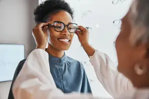 A smiling woman wearing glasses with a blurry background of an optometrist adjusting them on her face