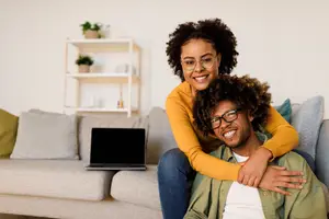 A man and a woman sitting on a couch, both wearing glasses and smiling, with a laptop on the couch behind them.