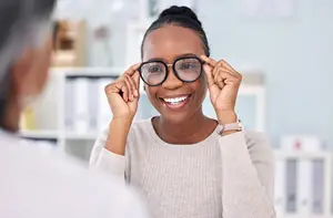 A smiling woman adjusts her glasses in an office setting.