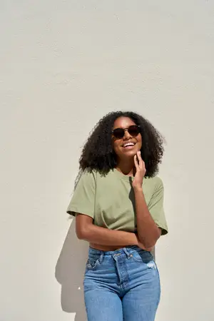 A smiling woman wearing sunglasses and standing against a white wall with her hands on her hips.