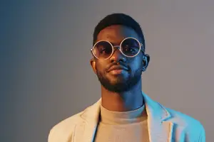 A black man in round glasses and a light blue coat smiles at the camera