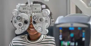 A girl wearing a striped shirt is having her eyes examined by an eye doctor using a machine with various dials and settings.